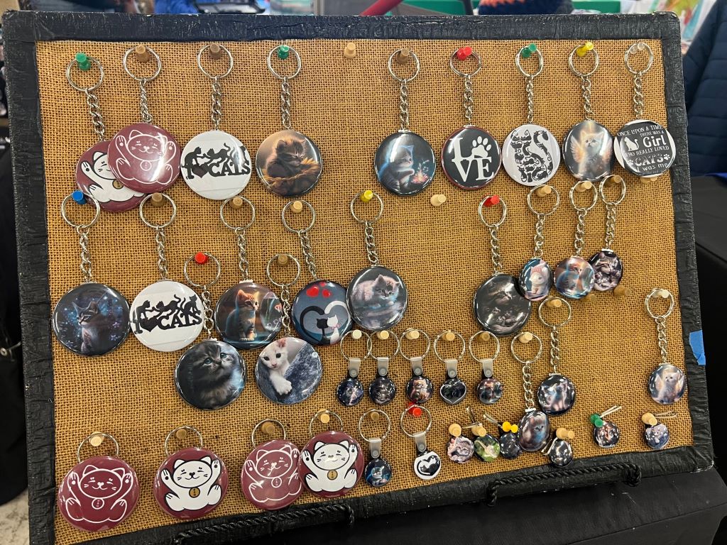 A board covered in burlap with push pins stuck in it. Keychains are hanging on each pin, and they are buttons with images of cats on them.