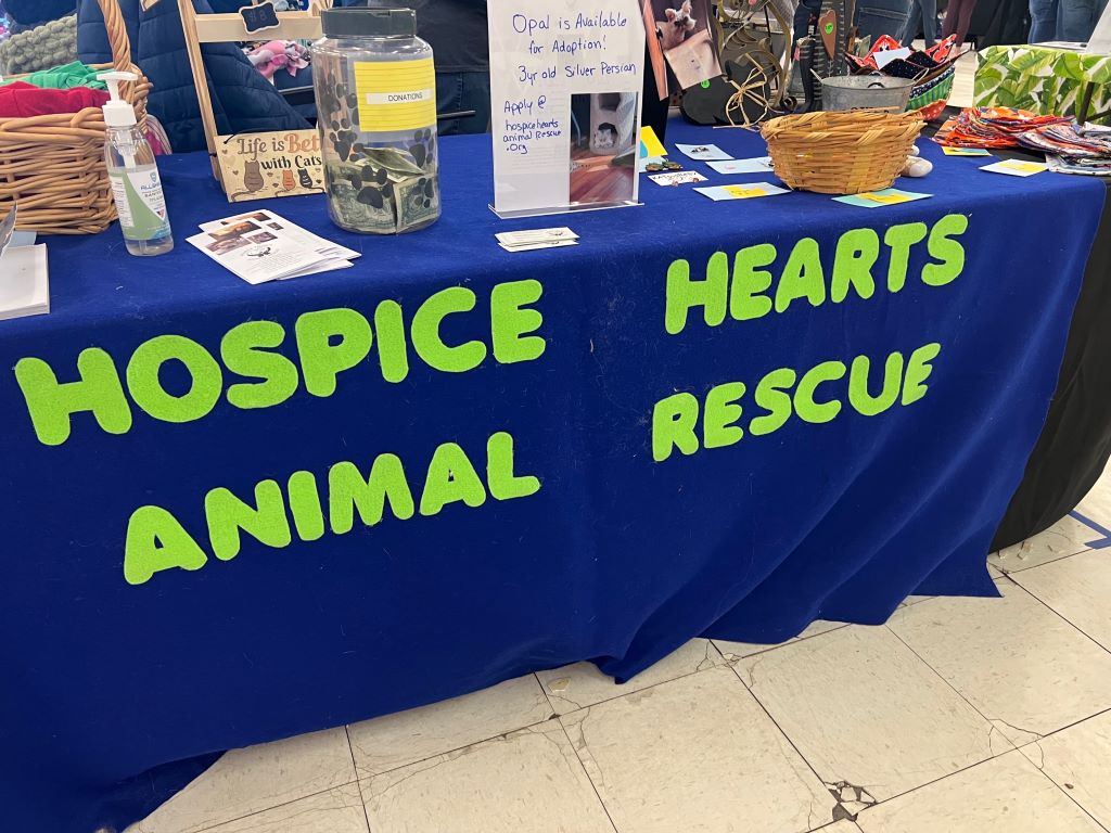 A table covered in a blue fabric tablecloth. It has Hospice Hearts Animal Rescue in lime green letters. There are various items displayed on the table.