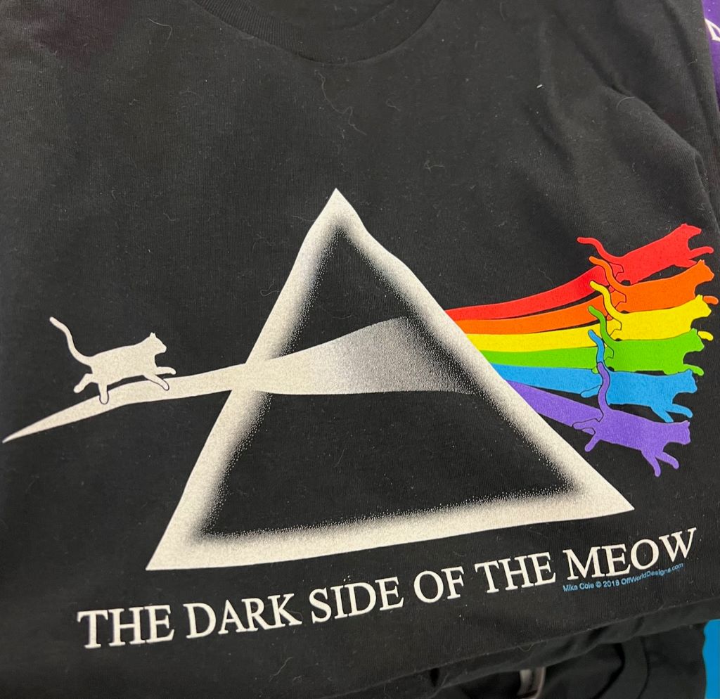 A close up of a black t-shirt with a white triangle in the center. The silhouette of a white cat is walking toward the triangle, and silhouettes of cats of each color of the rainbow are coming out the other side. In white letters it says The Dark Side of the Meow.