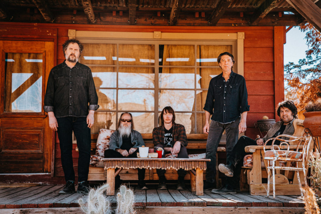 The band Drive-By Truckers are standing and seated on the porch of a wood house. They are five white-presenting men. Two are standing, two are seating on a couch, and one is seated in a chair.