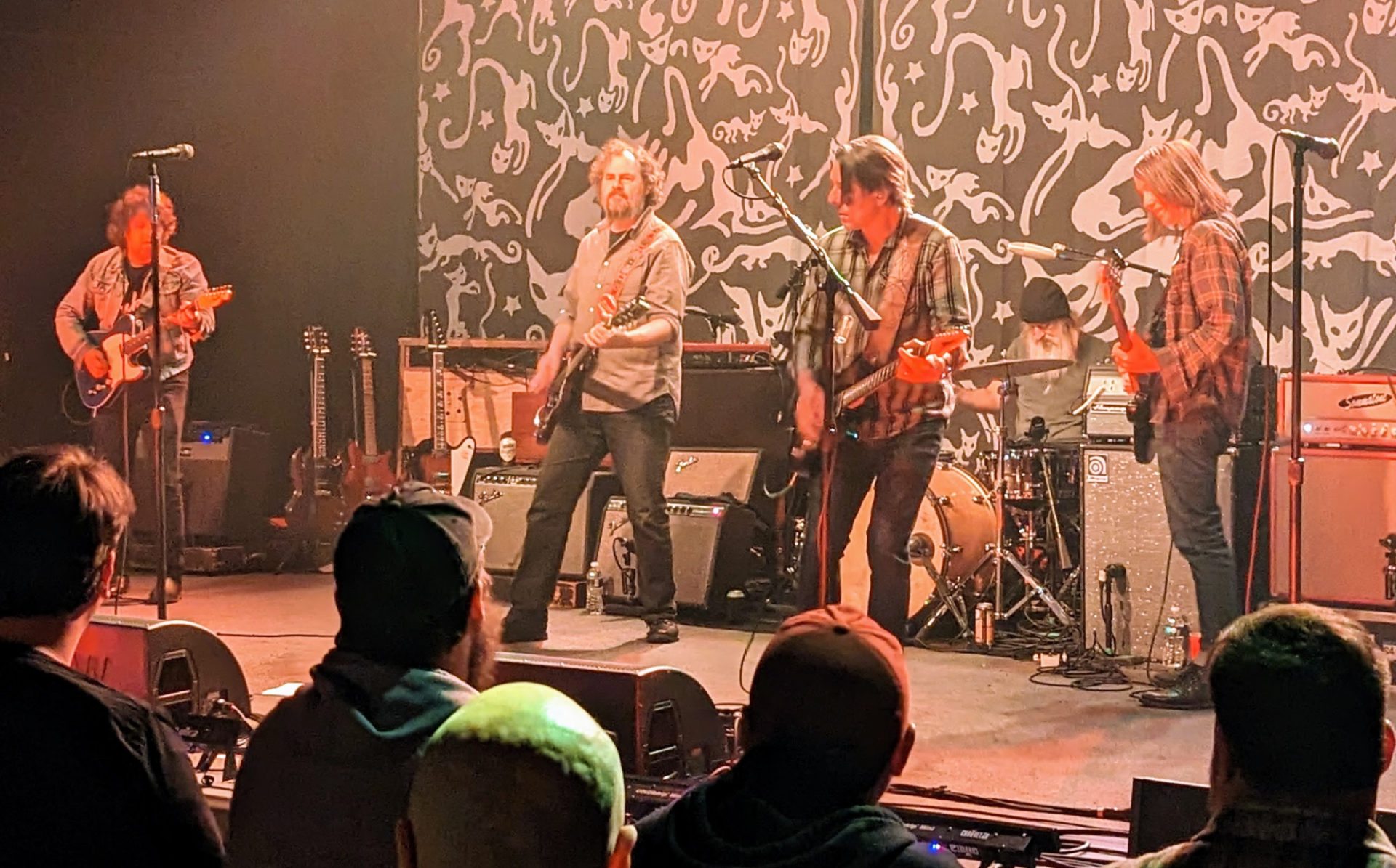 Drive-By Truckers performing at Canopy Club in Urbana. Five middle aged white-presenting men are playing instruments on a stage. A crowd looks on.