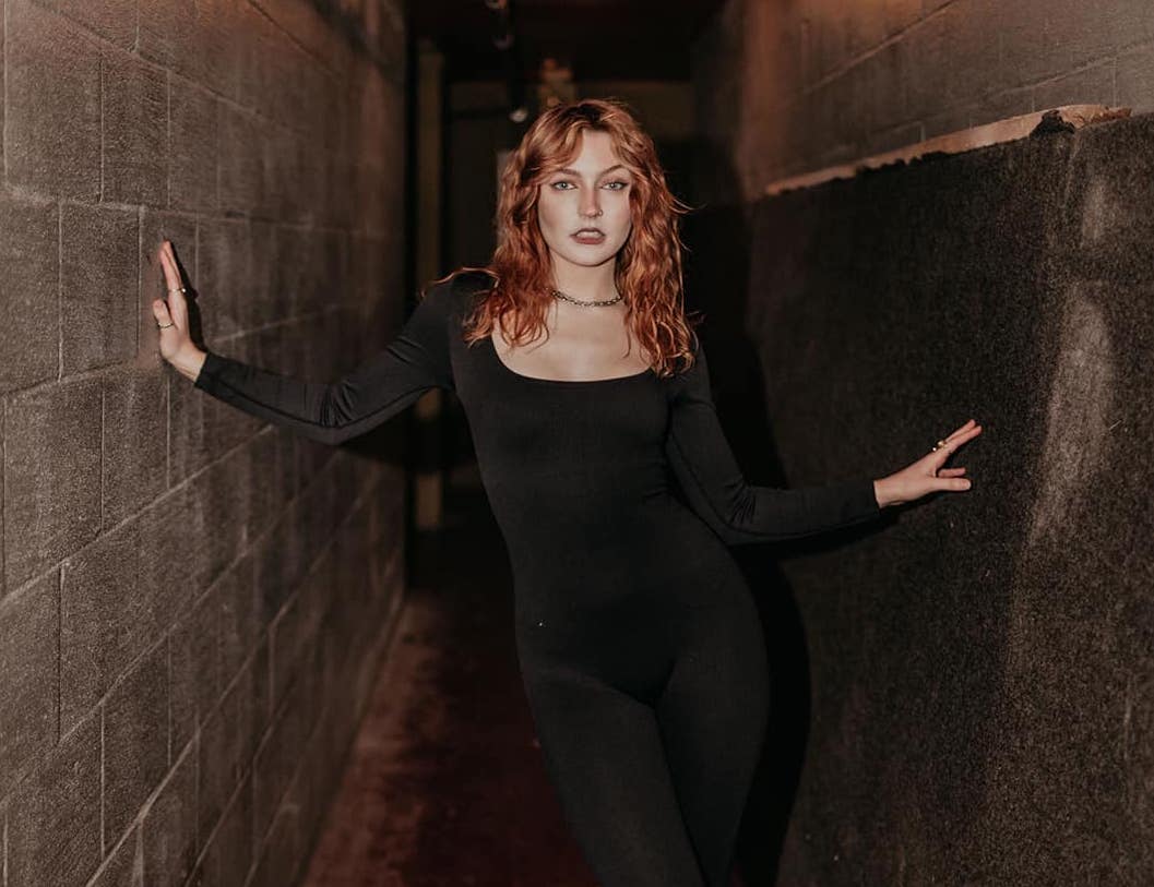 Musician Emily How photographed straight on. She is a tall, thin white woman with reddish hair. She is wearing a black, full body leotard and standing in a very narrow industrial hallway. She is touching her hands to each of the concrete walls.