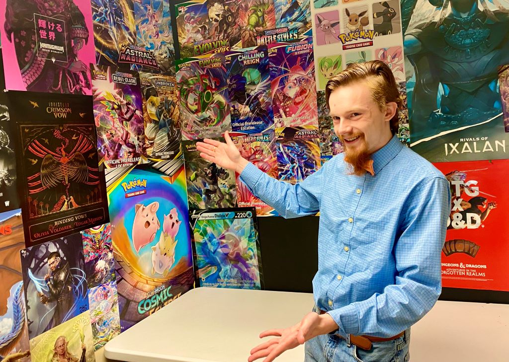 A white man with red hair and a red goatee, blue collared shirt and orange bow tie, is gesturing towards a wall covered in colorful game posters.
