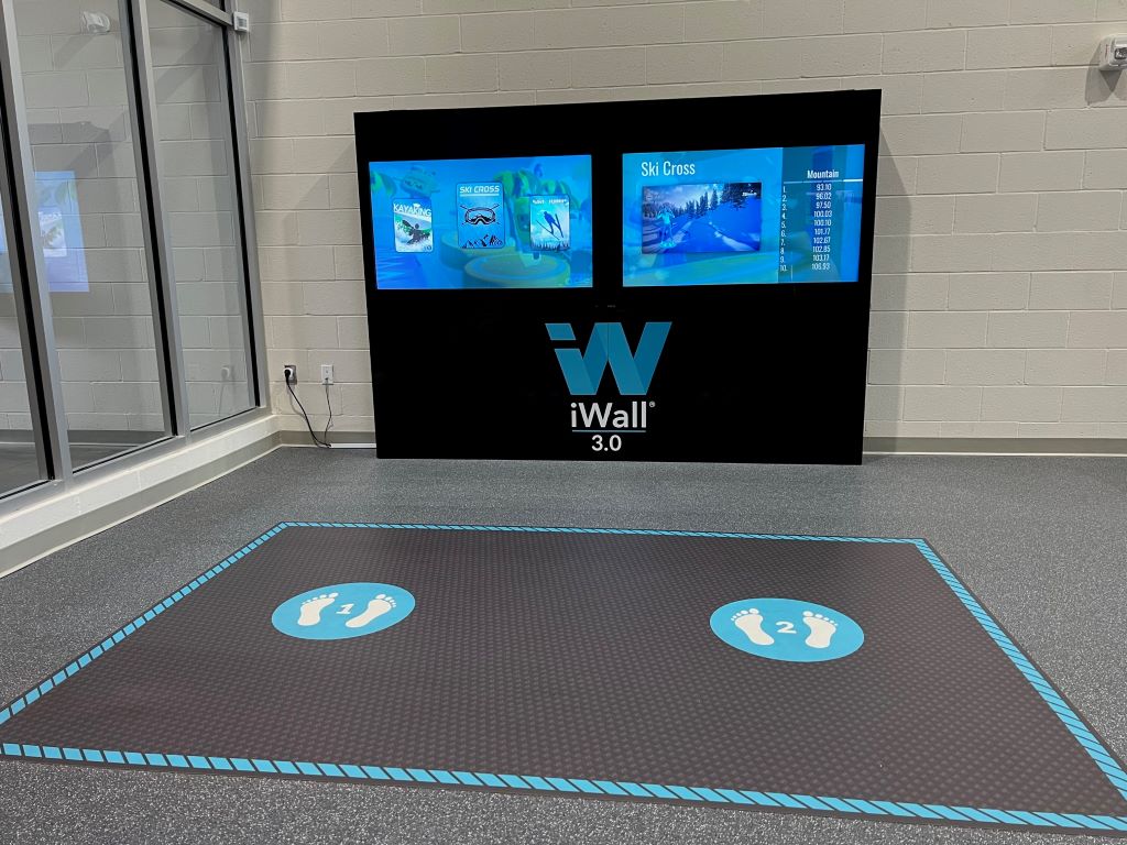 A console with two large tv screens is against a white concrete wall. There is a mat with two blue circles on the floor in front of the tvs. 