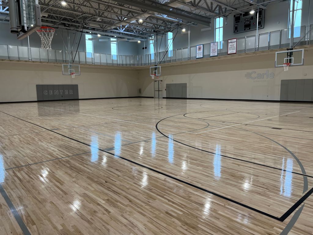 A large gym with shiny wood floors and basketball court markings. There is a walking track above the court, around the perimeter.
