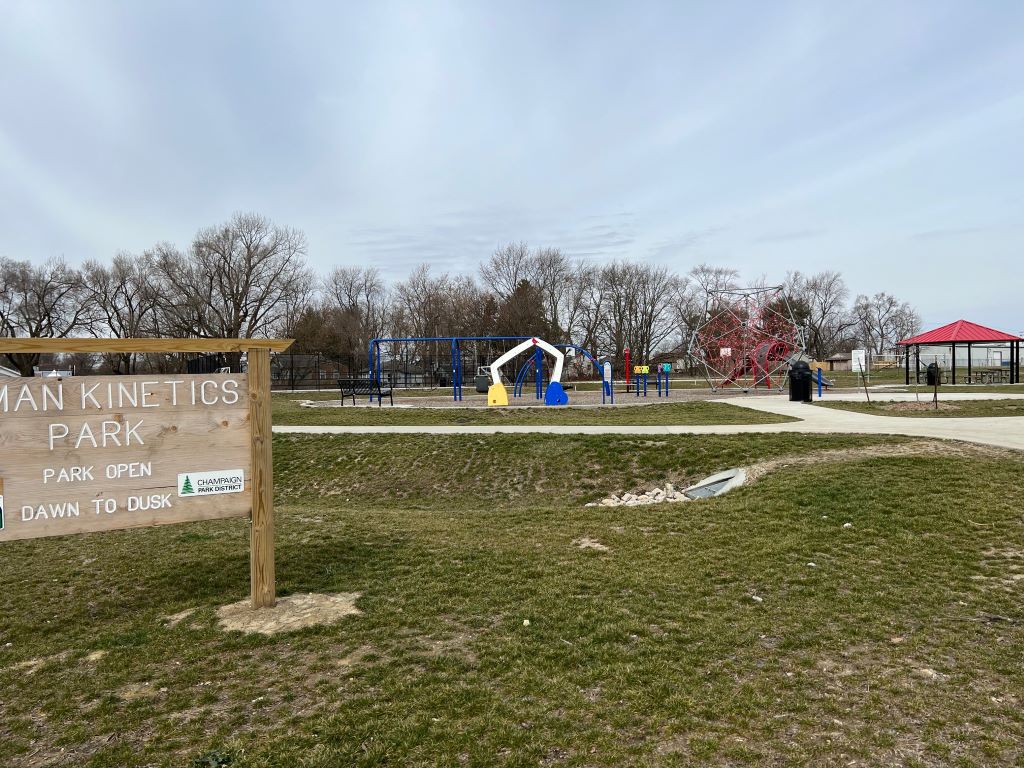A park with a playground, pavilion, and walking path. In the foreground is a wooden sign that says Human Kinetics Park in white lettering.