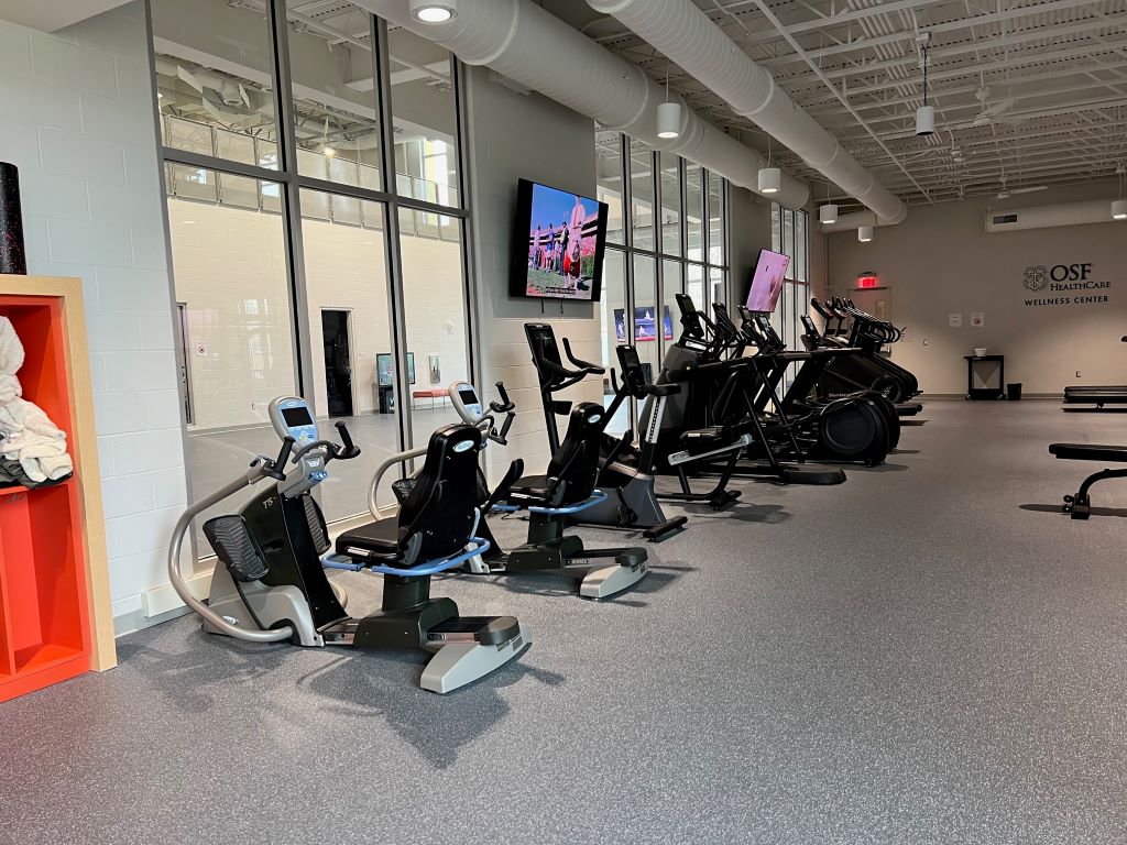 A row of elliptical machines and exercise bikes in a large room, with two televisions hanging above them.