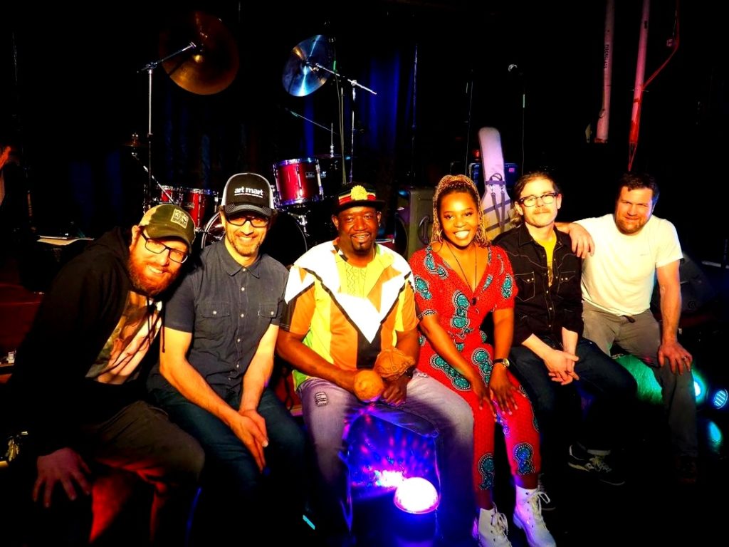 Members of the band Mhondo Rhythm Success sitting down on a stage, looking at the camera and smiling.