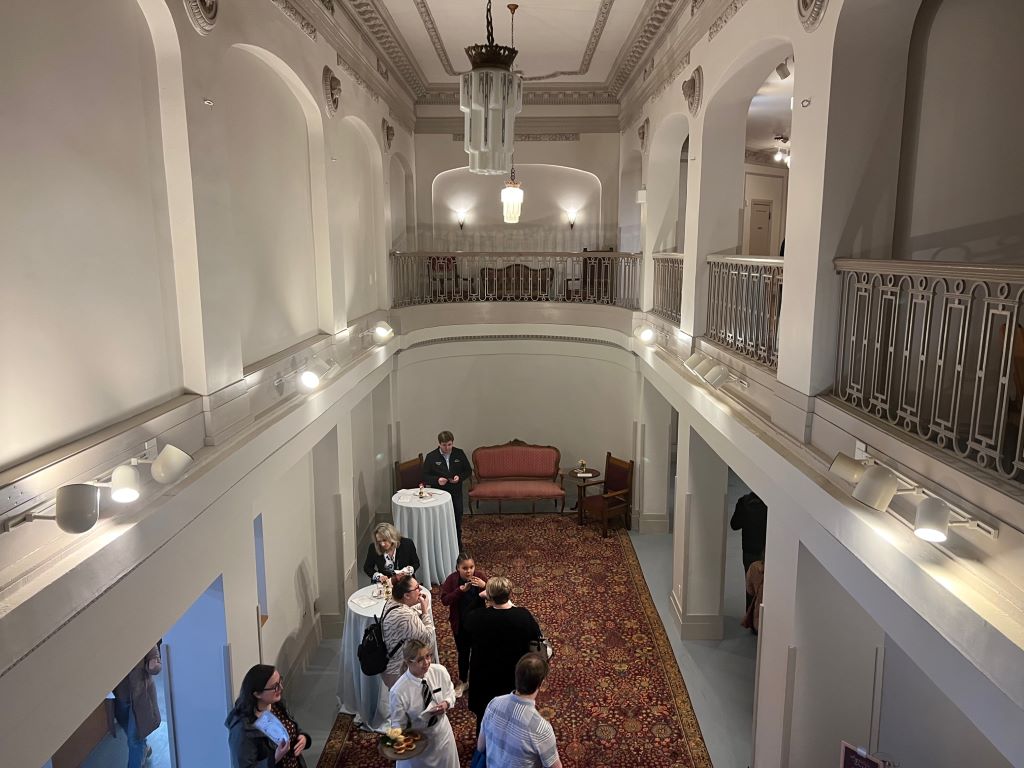 A view from the second floor of the Orpheum Theatre. The walls are white, and the room is cylindrical. There is a railing along the second floor, and you can see down to a lower level with high top tables and people milling around.