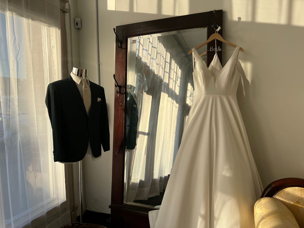 A wedding dress is hanging on a large full length mirror. A black suit jacket is on a stand up form on the other side of the mirror.