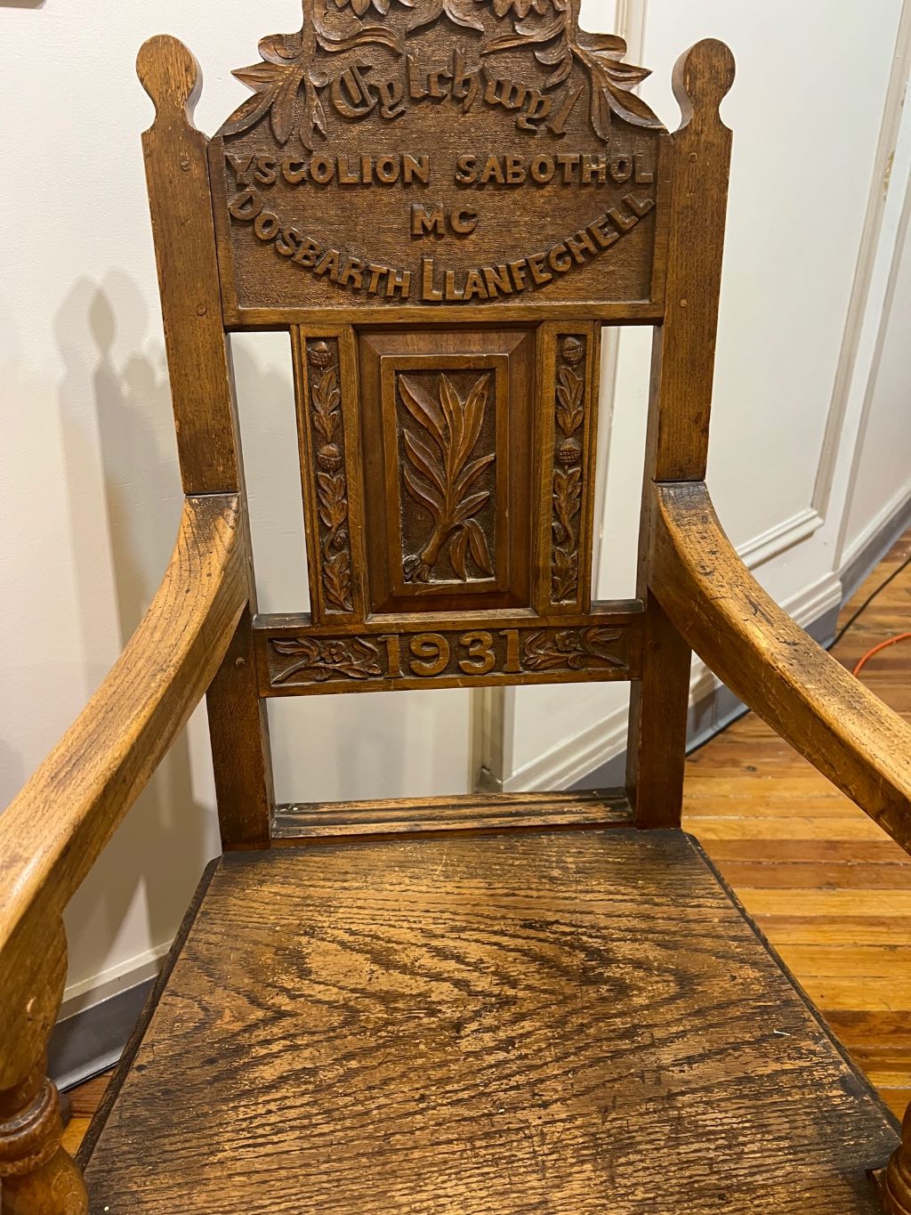 A wooden chair with carvings of leaves and flowers and words in Welsh.