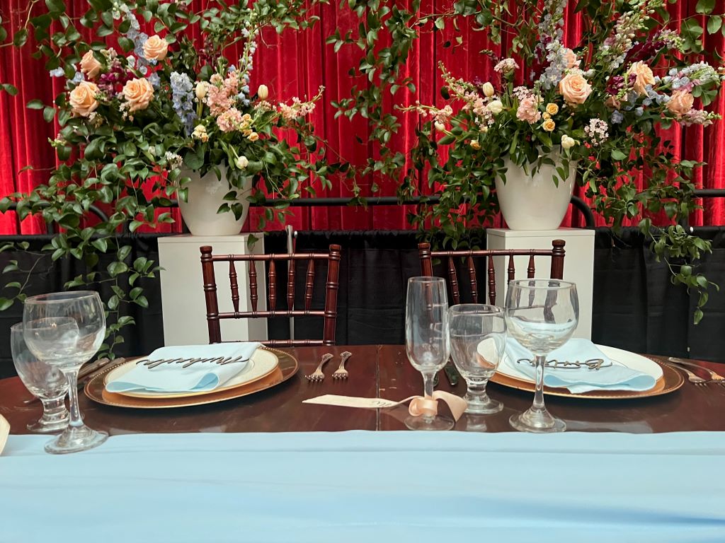 A table with a light blue tablecloth and two place settings. There are two large floral arrangements in white pots on top of white pedestals. 