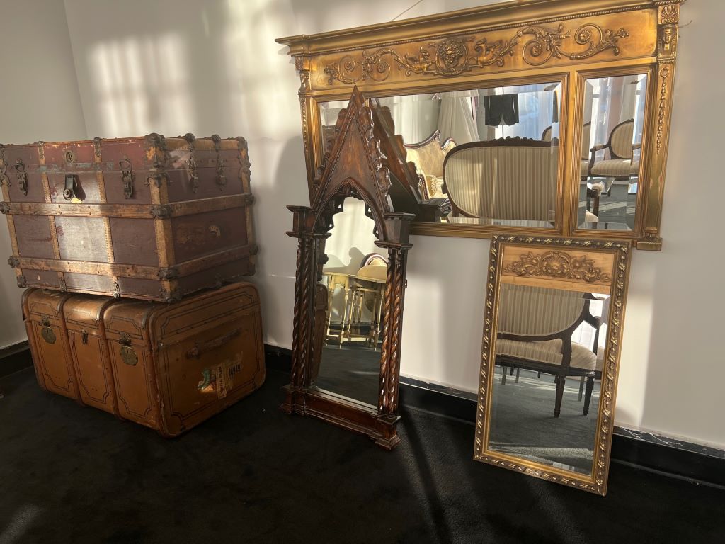 Two vintage brown trunks are stacked on top of each other. There are a couple of decorative mirrors leaning against the wall, and a large mirror hanging on the wall. 