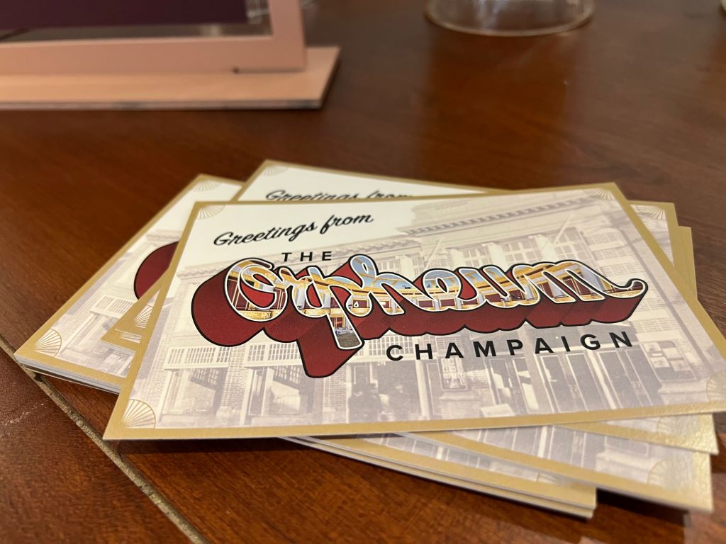 A stack of postcards on a dark wood tables. The face of the postcard says "Greetings from The Orpheum Champaign"