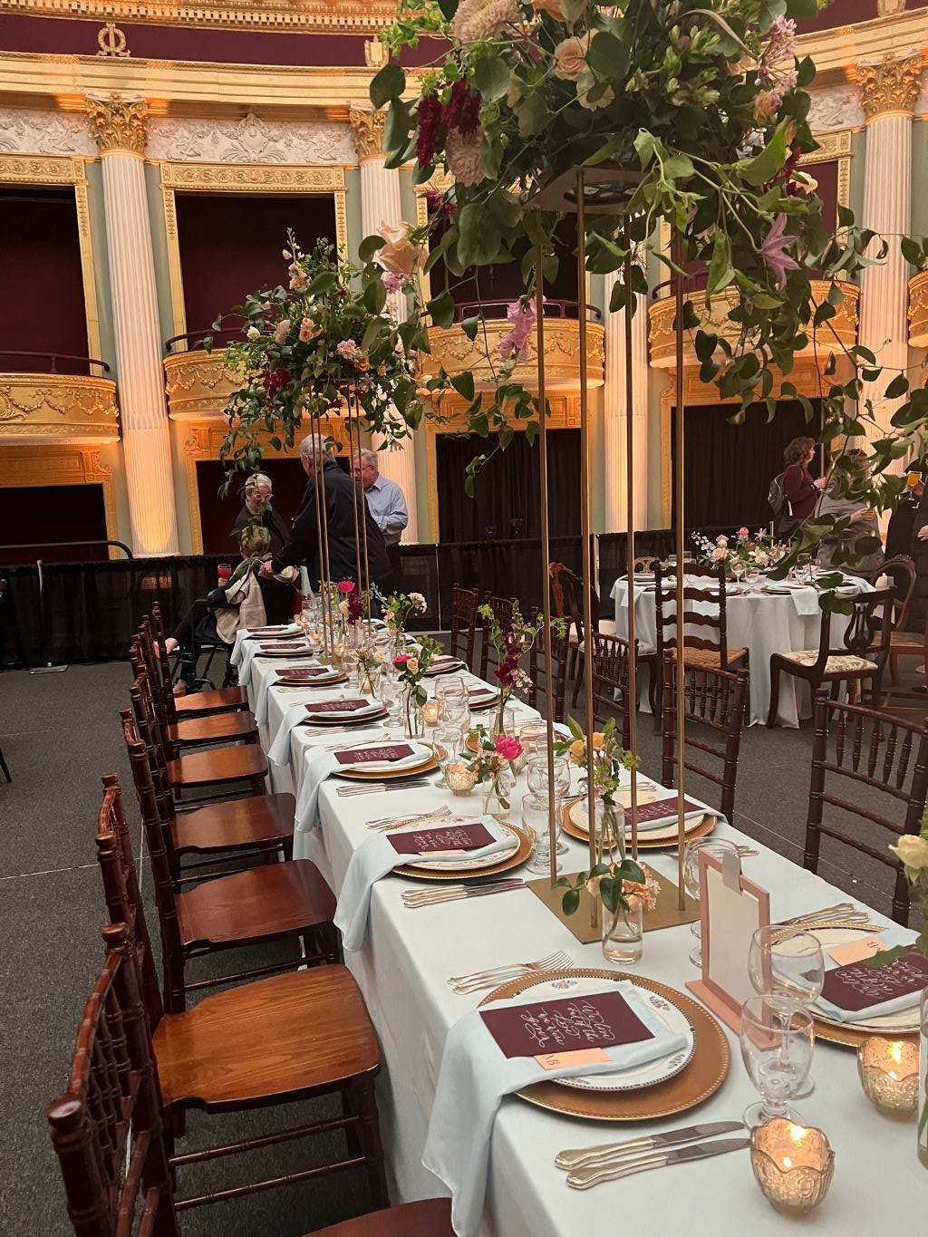 A long table with a white tablecloth and brown wood chairs. There are place settings in front of each chair, and tall floral arrangements. 