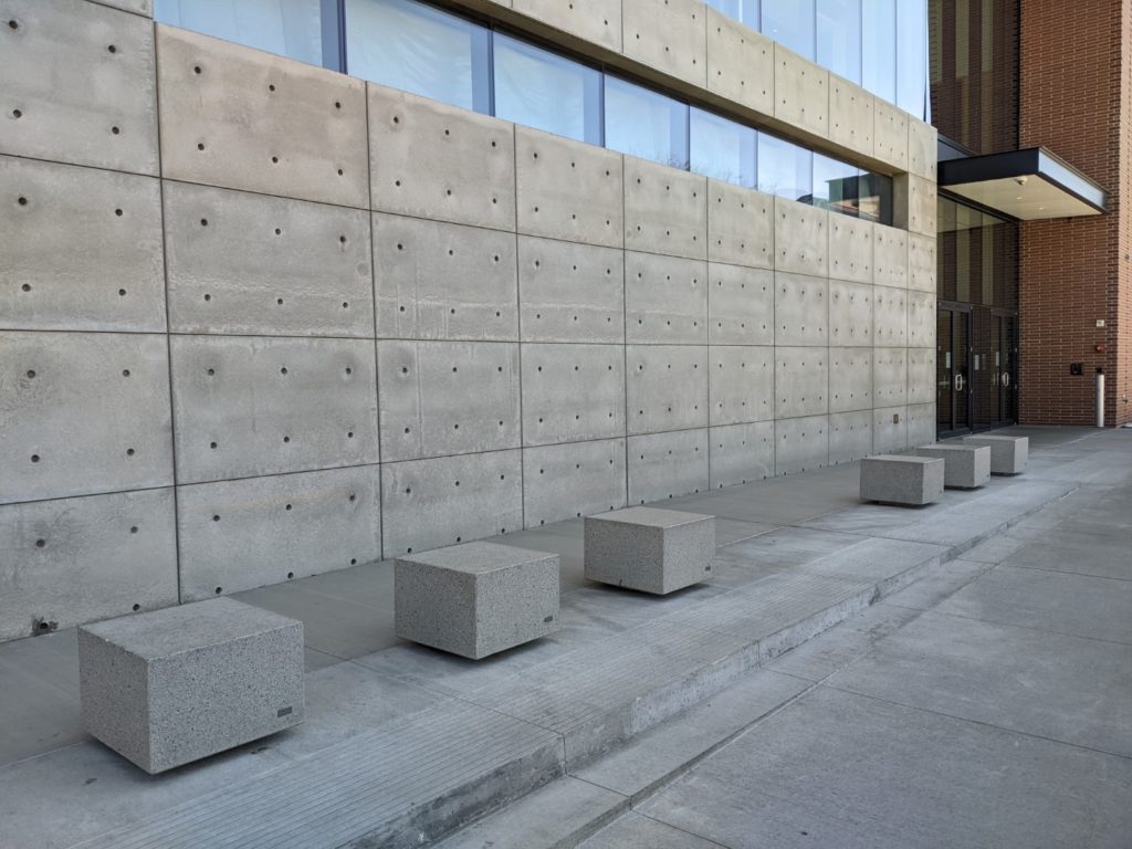 A gray stone wall that is sectioned into rectangles. Six gray stone cubes sit on the sidewalk in front of it.