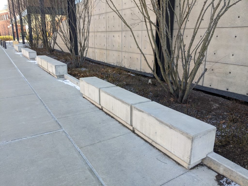 Long rectangular stone benches are placed along a sidewalk. Behind them is a space with dirt and several small trees.