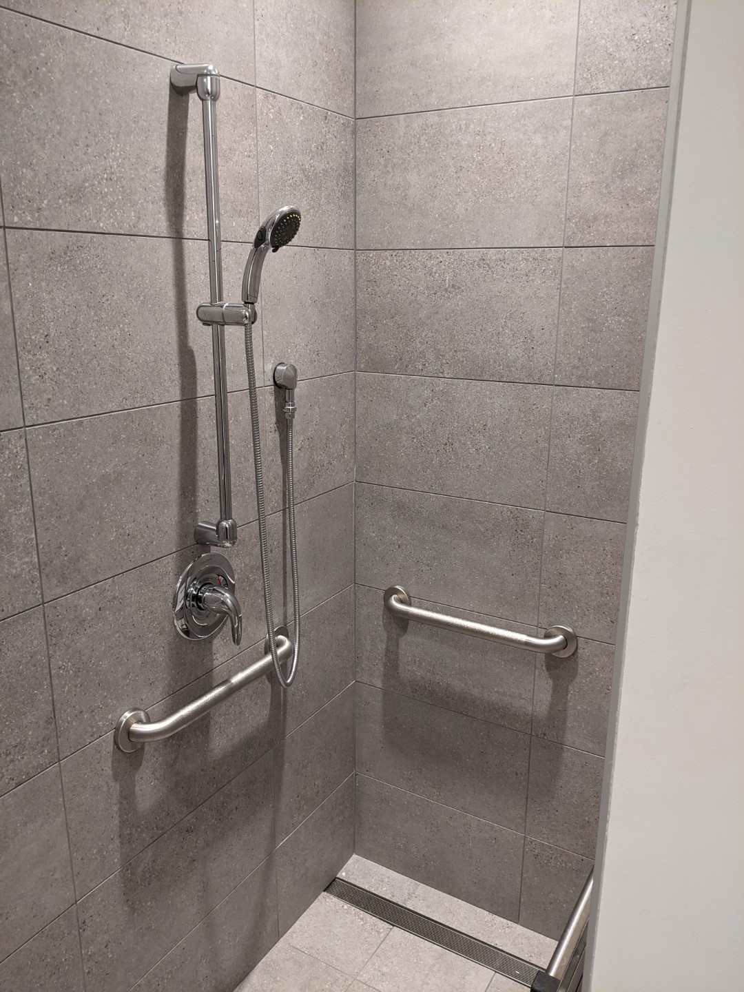 A metal shower head and faucet is attached to a gray wall. There are metal hand rails attached to each wall as well.