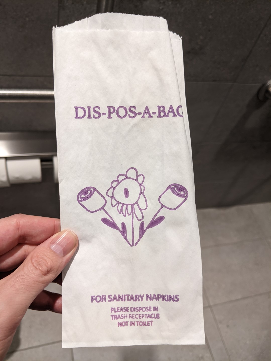 Close up of a paper bag with purple printing on it. It says Dis-pos-a-bag and has an image of a flower and two rolls of toilet paper on stems.