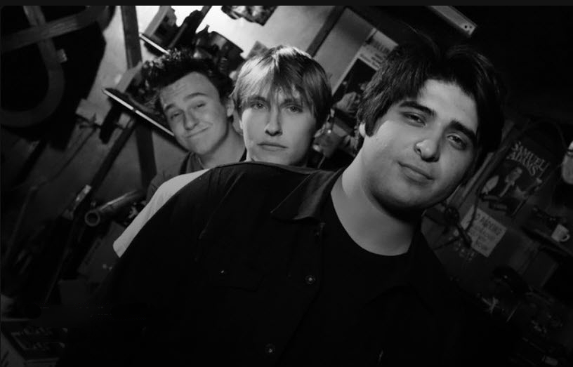 A black and white photo of the band Pure Intention. Thee young men are pictured in a line with their heads titled so their faces are visible. They all appear to be white or white passing. The three of them have shaggy hair and mostly neutral or slightly smirking expressions. They are standing in what appears to be a bar or basement space. 