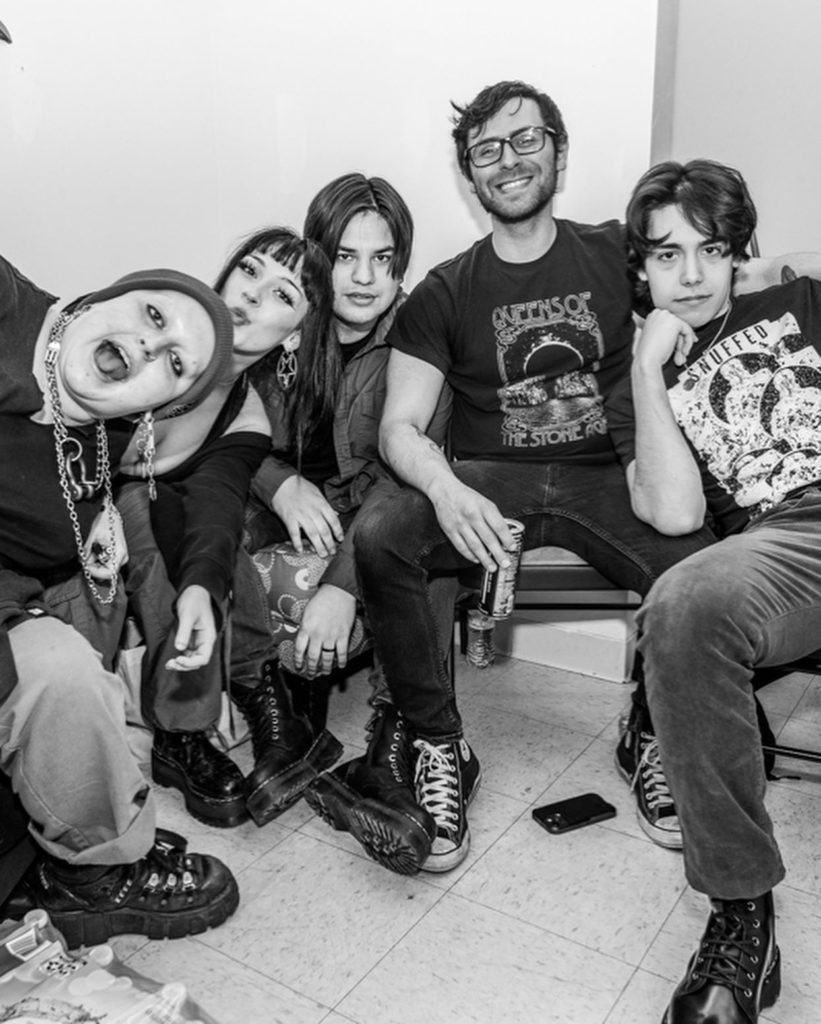 Black and white photo of the band Scarlet Demore. There are five people, all dressed in black tshirts and jeans. They are posing for a photo, some of them making funny faces. 