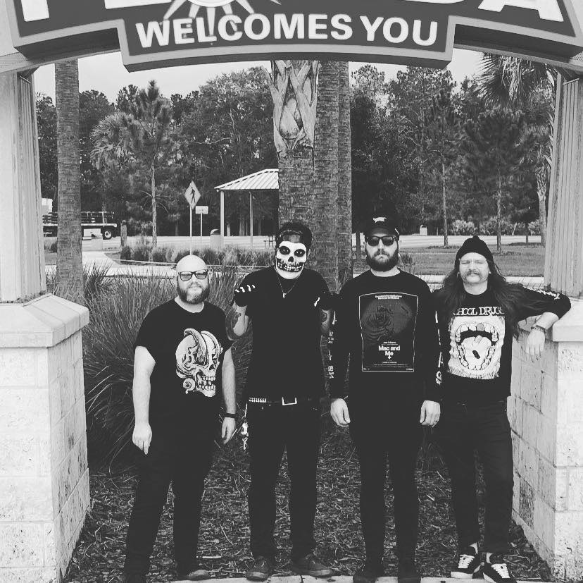 Black and white photo of all four band members standing underneath a sign that says "Welcomes You".