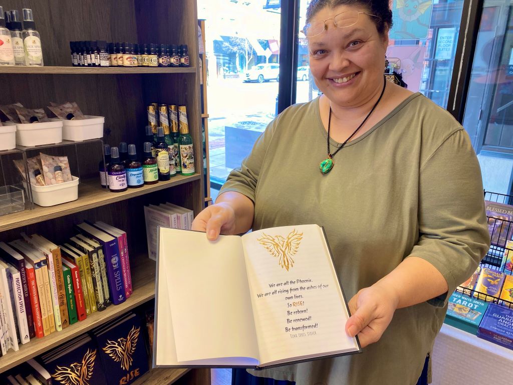 A woman with an sage green shirt, a bright green pendant, and dark hair is holding a book open to a page with black script and a gold emblem of a hawk.