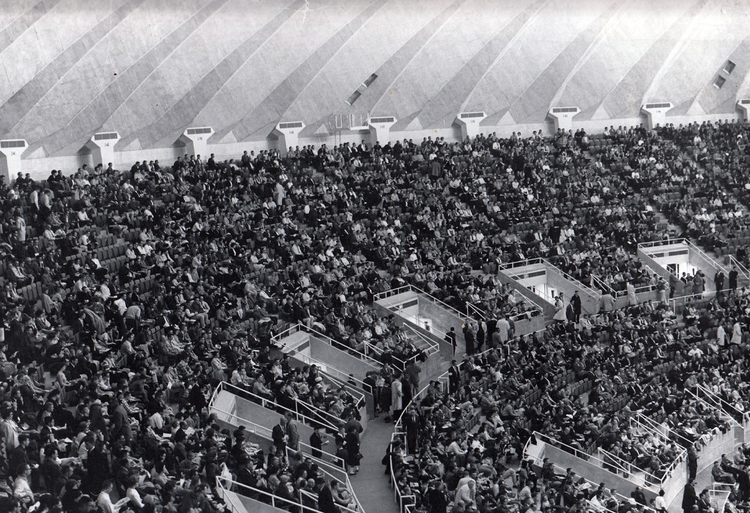 A black and white photo of a portion of an arena, with people filling the seats and a portion of a white domed roof.