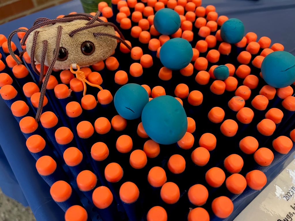 A bunch of orange tipped nerf darts are set up in a grid. There are blue balls of clay sitting on top of them. There is also a small brown fuzzy stuffed spider. 