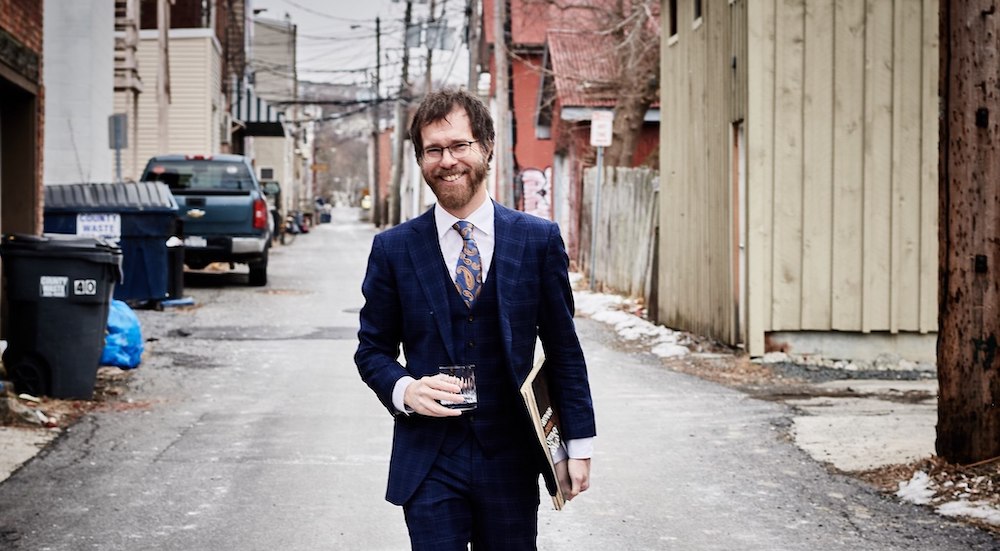Ben Folds stands in an alleyway, smiling, holding a drink in his right hand and what looks to be a few 12 inch records in his left hand.