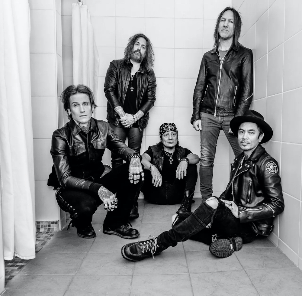 A black and white photo of the five members of Buckcherry. They pose in a bathroom, most of them in leather jackets and black pants. They look into the camera. 