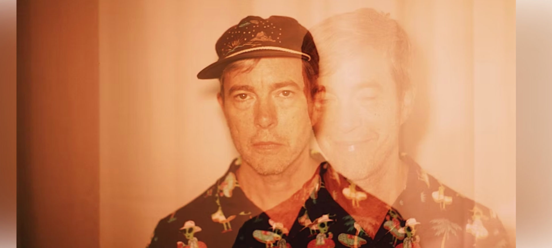 Bill Callahan portrait. He is pictured wearing a dark collared shirt with a bold pattern and a baseball hat. He looks into the camera with a blank expression. There is a ghost image effect that has a faint outline of his face to the right of the in-focus face. That ghost image shows him without a hat, eyes closed, smirking. The image is filtered in a pale orange.