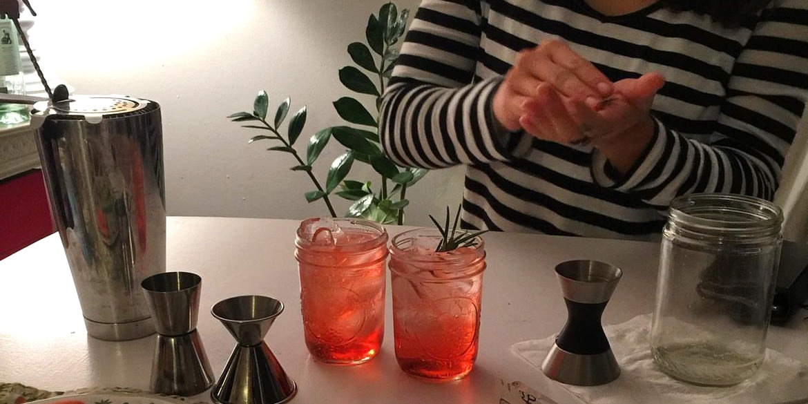 On a white counter, there are two glasses with a reddish mocktail. The bartender has a black and white shirt and is cupping their hands together. Photo from Katie's Cocktails on Instagram.