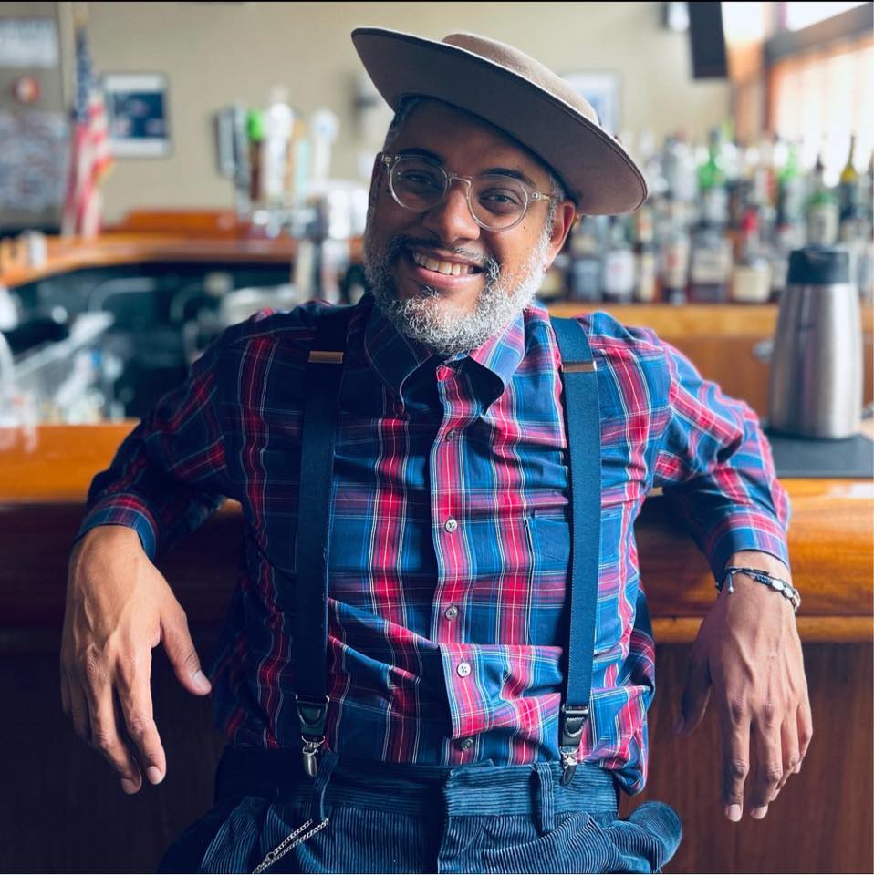 Dom Flemons sits at a bar, facing the camera and smiling. He wears a blue and red plaid shirt with blue suspenders and a tilted boater hat.