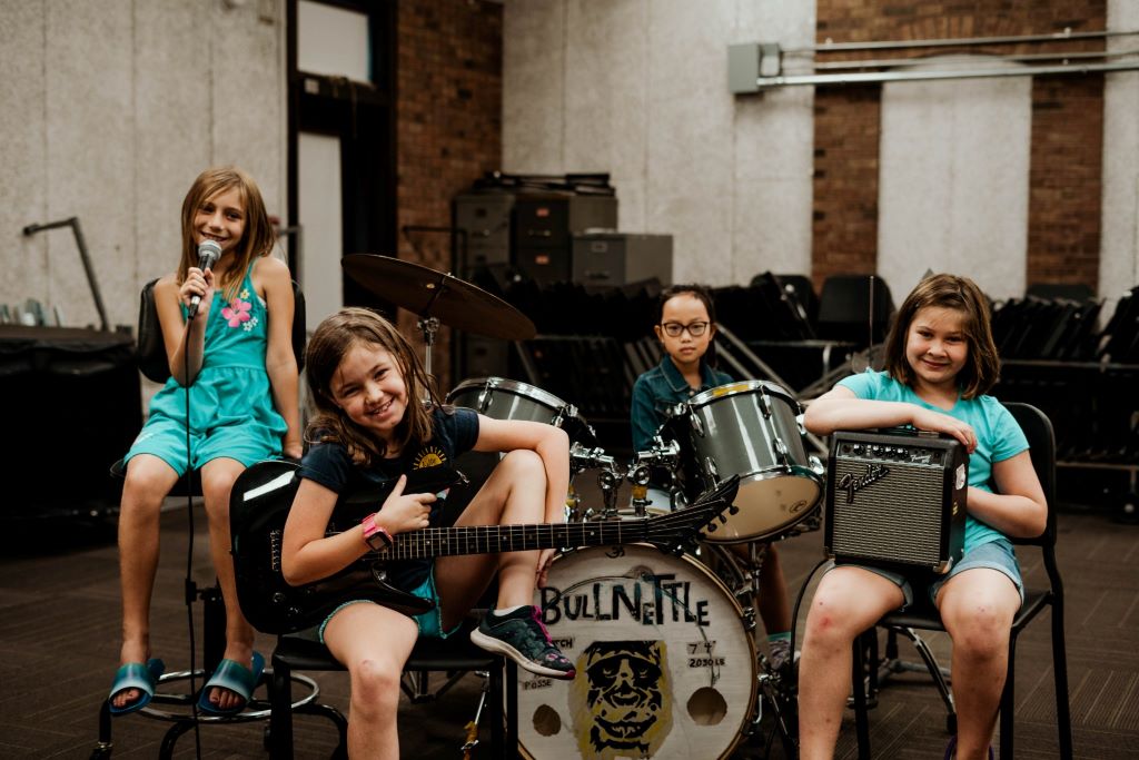 Four young girls are gathered around a drum set. One is holding a microphone, one is holding a bass guitar, and one is holding an amp.
