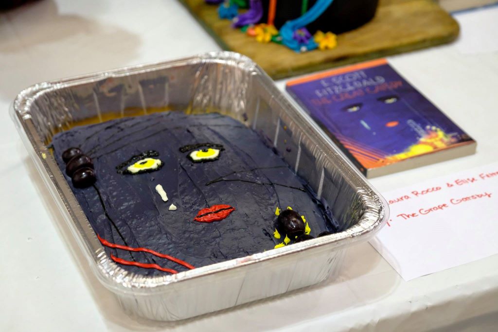 An aluminum cake pan is filled with a cake with dark purple icing, yellow eyes, and red lips. It is sitting next to the book The Great Gatsby.