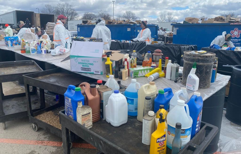 A long table and a rolling cart have various sizes of bottles of chemicals sitting on them. There are people in white hazmat suits standing behind the table.