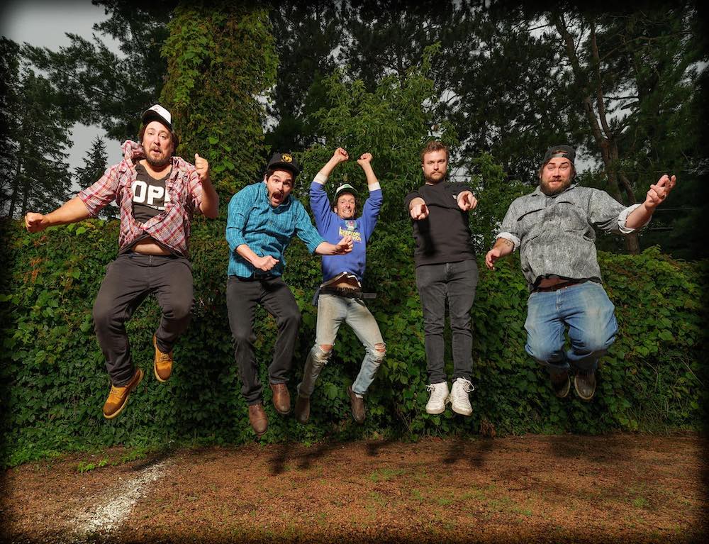 The 5 members of Horseshoes and Hand Grenades are caught jumping into the air. They are outside in front of what looks to be a forest. 