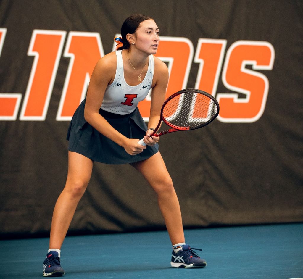 An Asian woman is wearing a white tank and dark blue skirt. She is crouched and holding a tennis racket in front of a sign that says Illinois in orange block letters.