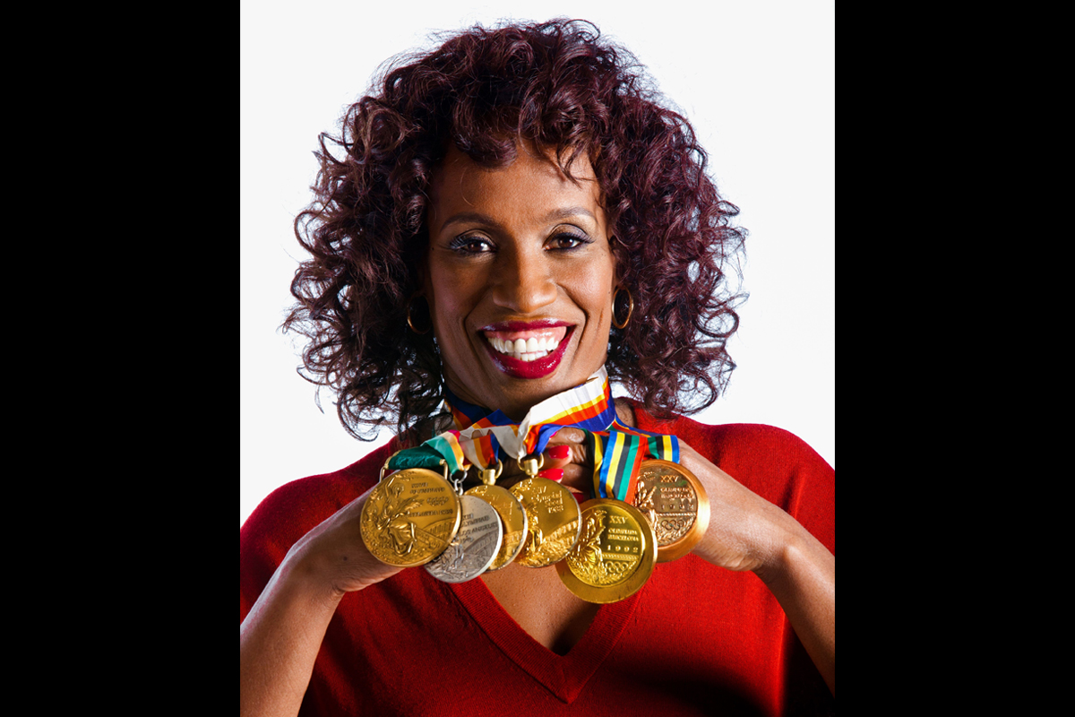 A Black woman with curly dark brown hair has gold and silver medals around her neck that she is lifting to show the camera.