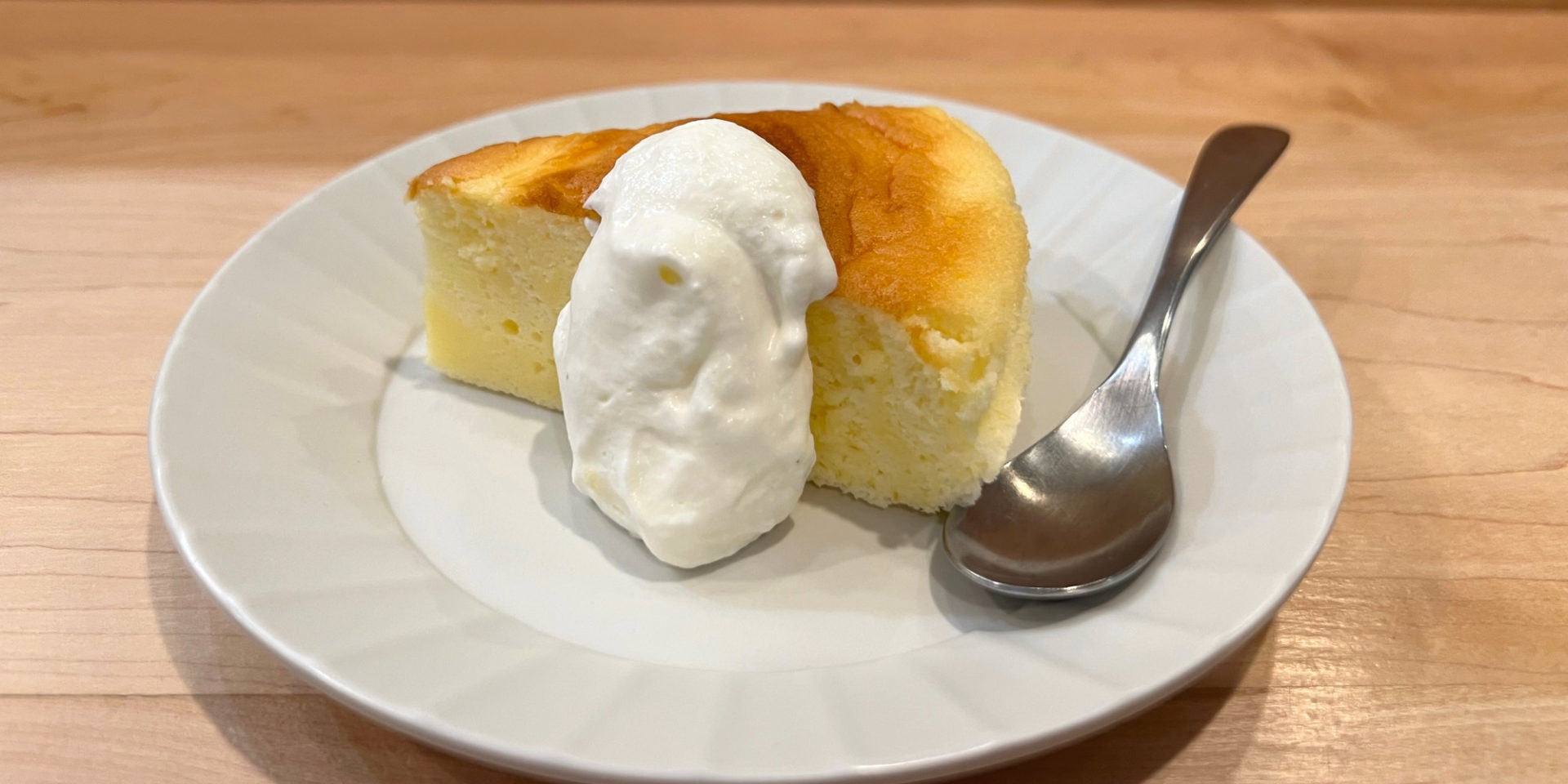 A slice of Japanese cheesecake by Kaori's Oven sits on a white plate with a dollop of fresh whipped cream and a metal spoon. Photo by Alyssa Buckley.