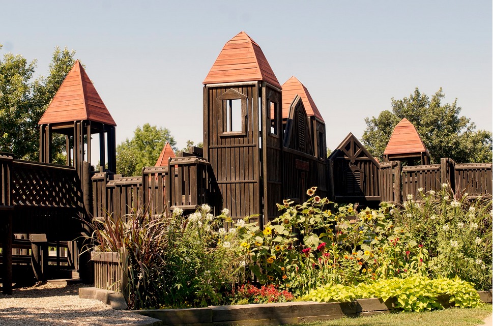 A playground made of dark brown wood with orange pointed roofs. There is an area of wildflowers in front of it.