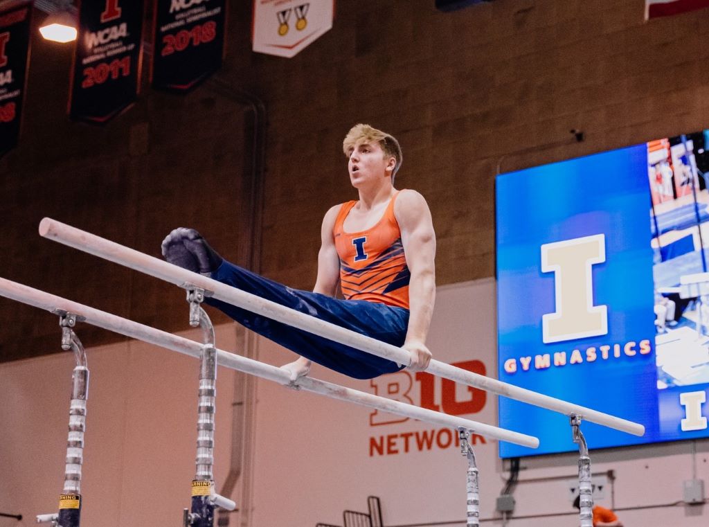 A man is propped on parallel bars with his legs straight out in a pike. He is wearing an orange tank and blue pants. 
