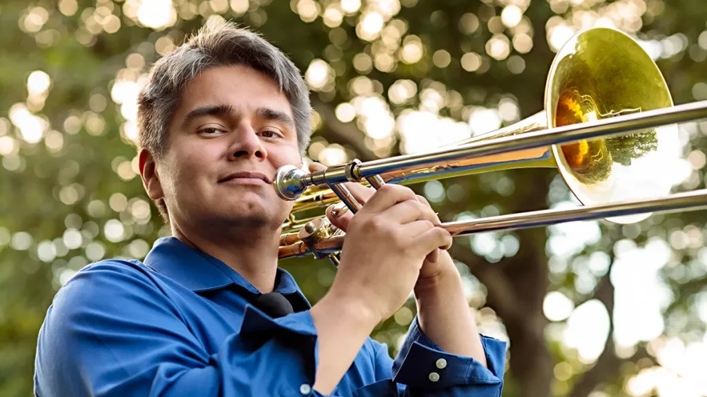 Peter Tijerina pictured from the shoulders up. He is wearing a blue collared shirt and holding a trombone just in front of his mouth as if he is about to play it. He looks into the camera. The background is  out of focus trees. 