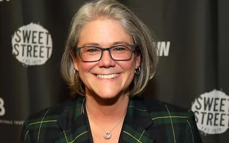 A white woman with short grayish hair and glasses. She is smiling, and is wearing a green plaid jacket.