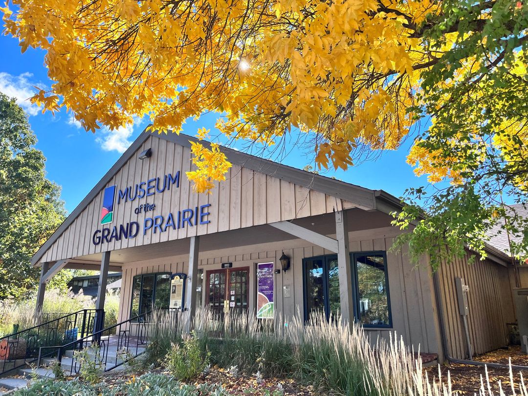 The front of a one story building with beige vertical siding and a pointed roof. It says Museum of the Grand Prairie in blue lettering. There are trees with yellow and green leaves surrounding it.