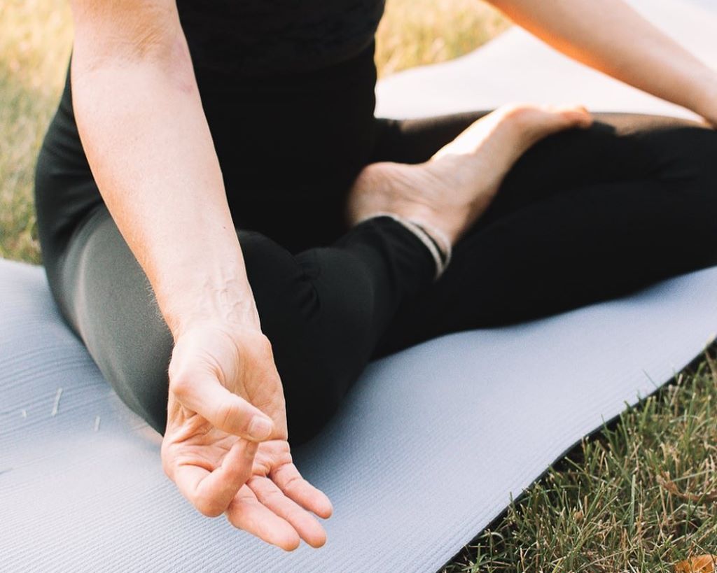 Close up of a person in black leggings and a black top sitting cross-legged with their arms outstretched and resting on their knees. They are sitting on a yoga mat in the grass.