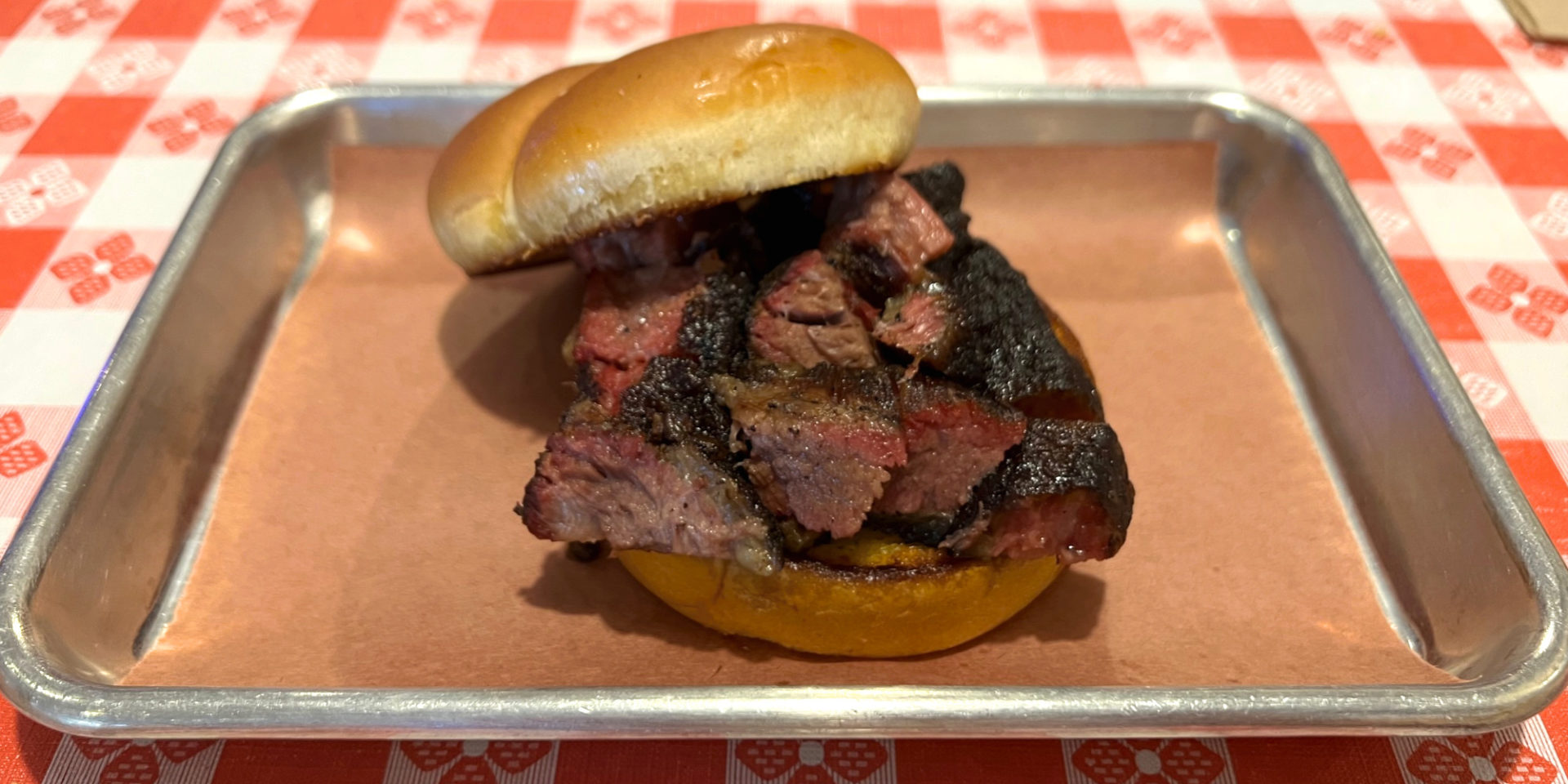A burnt ends sandwich from Smoky's House BBQ sits on a metal tray atop a red-and-white checkered tableclothed table. Photo by Alyssa Buckley.