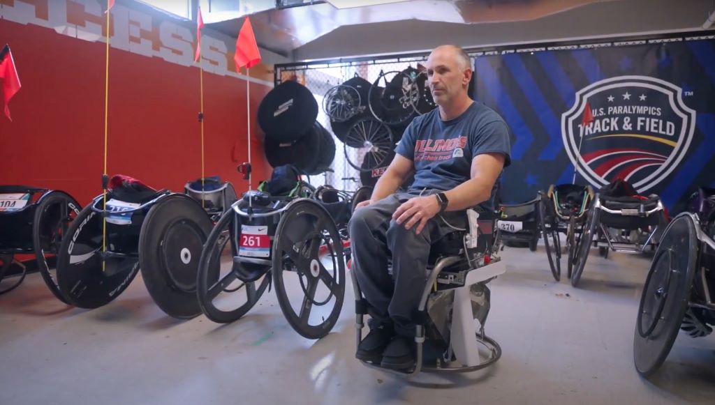 A man is sitting in a robotic wheelchair, with other wheelchairs sitting nearby. He is wearing a blue t-shirt and jeans.