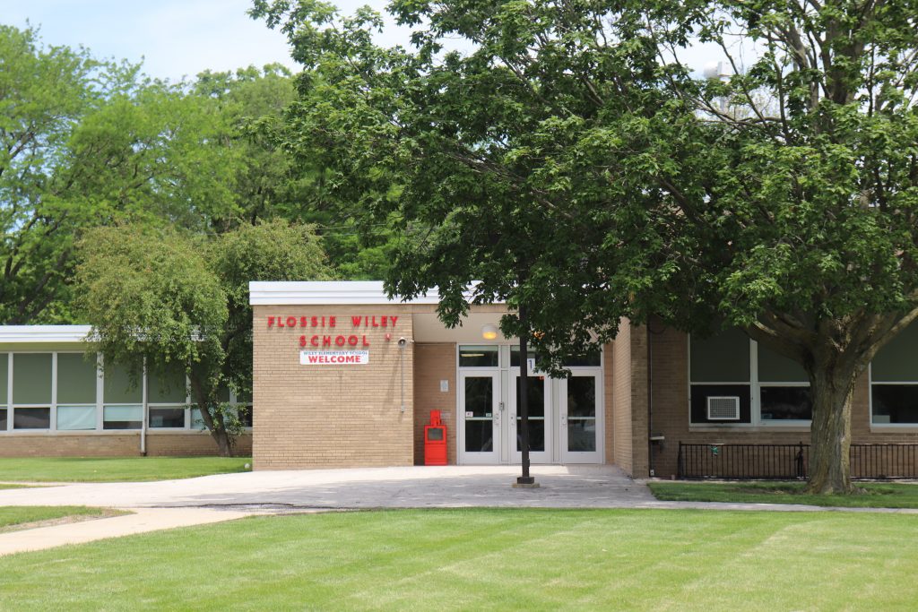 The facade of a light brown brick school building with three glass doors. It saw Flossie Wiley Elementary in red letters. There are leafy green trees surrounding the building, and a grassy area in front of the entrance.